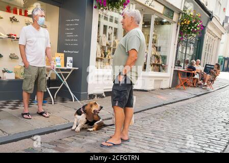 Canterbury, Regno Unito - 13 agosto 2020 un uomo che indossa una maschera a causa di Covid 19 e che tiene un cane Basset Hound su un capo hanno una conversazione in piedi su un quai Foto Stock