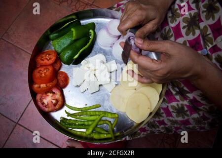 Donna indiana che trita le verdure con entrambe le mani usando il coltello. Taglio di verdure indiane in acciaio inox thaali. Le attività domestiche quotidiane Foto Stock