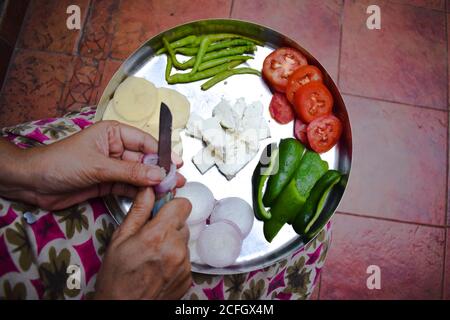 Donna indiana che trita cipolle con un coltello con entrambe le mani su Thali. Preparazione per fare pakoda o curry sabji su thali. Metodo tradizionale o Foto Stock