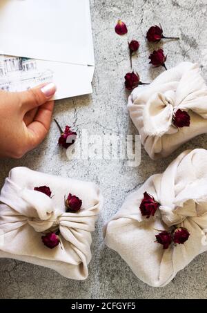 Regali imballati in furoshiki, rose secche e mani che ottengono una cartolina da una busta su uno sfondo grigio. Foto Stock