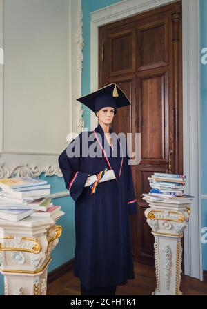 Manichino studentesco, all'interno di una biblioteca, vestito da laureato con un cappello nero accademico e abito, tenendo un diploma in mano. Bella ornata antico pil Foto Stock