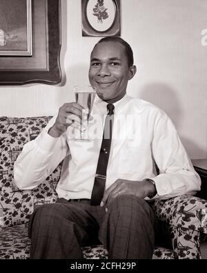 1960 UOMO AFROAMERICANO SORRIDENTE SEDUTO SUL DIVANO BEVENDO VETRO PILSNER BIRRA CHE INDOSSA MANICHE SHIRTSLEEVES CRAVATTA GUARDANDO LA FOTOCAMERA - N1830 HAR001 HARS PERSONE MASCHI FIDUCIA B&W OCCHIO CONTATTO FELICITÀ BEVANDA ALLEGRA LEISURE AFRO-AMERICANI AFRO-AMERICAN FLUID RELAXD NERO ETNIA ORGOGLIO NICKTIE SORRIDE PILSNER CONSUMARE IDRATAZIONE GIOIOSO ELEGANTE SHIRTSLEEVES BREWSKI MID-ADULT MID-ADULT UOMO RINFRESCANTE RELAX A HOME BEVANDE BIANCO E NERO HAR001 AFROAMERICANI IN VECCHIO STILE Foto Stock