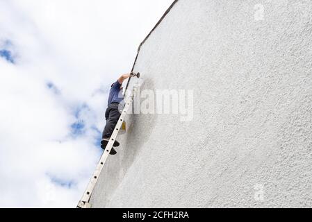 Uomo che lavora duro in alto una scala dipinge il lato della casa un colore bianco fresco. Foto Stock