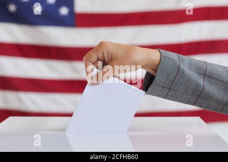 Vista laterale primo piano di una donna irriconoscibile che mette il bollettino di voto in una casella di voto mentre si è in piedi contro la bandiera americana il giorno delle elezioni, copia spazio Foto Stock