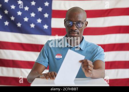 Ritratto di un uomo afroamericano moderno mettendo il bollettino di voto nella scatola del voto e guardando la macchina fotografica mentre si è in piedi contro la bandiera americana il giorno delle elezioni, copia spazio Foto Stock