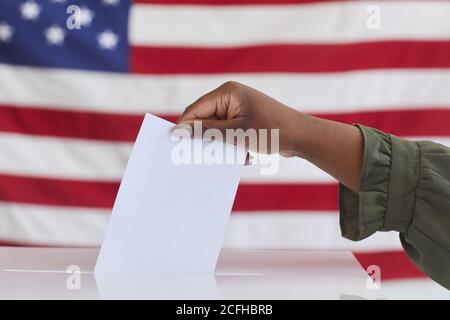 Vista laterale primo piano di una donna afroamericana irriconoscibile che mette il voto in una casella di voto contro bandiera americana sfondo, copia spazio Foto Stock