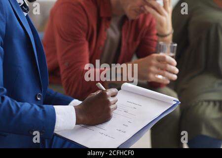 Primo piano dello psicologo afroamericano maschio che scrive sulla clipboard mentre guida sessione di gruppo di supporto, spazio di copia Foto Stock