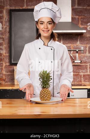 giovane chef cuoca in berretto bianco e uniforme con ananas sul piatto di fronte a lei Foto Stock