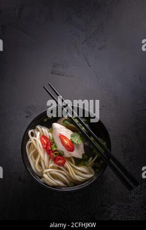 Piatto con piccante zuppa asiatica. Brodo di pollo e carne, peperoncino rosso, coriandolo e scalogno, tagliatelle giapponesi in ciotola nera con bastoncini Foto Stock
