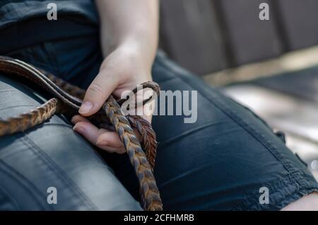 Primo piano della mano di una donna con guinzaglio in pelle. Una donna adulta tiene un guinzaglio in pelle di vimini per un animale domestico seduto su una panca del parco. Riposo durante una passeggiata. An Foto Stock