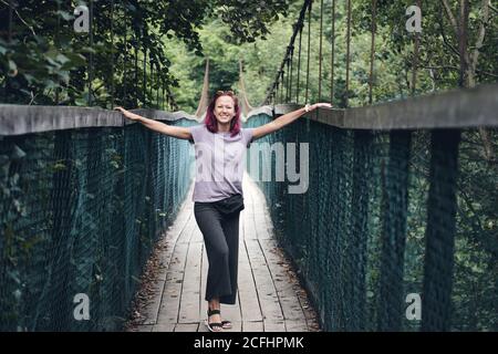 Giovane donna caucasica con capelli rosa sul ponte sospeso in acciaio lungo. Vecchio piccolo ponte di legno sul fiume solo per pedoni. Fiume di montagna Foto Stock