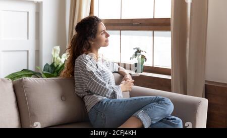 Bella donna pensiva che guarda fuori finestra mentre si riposa sul divano Foto Stock