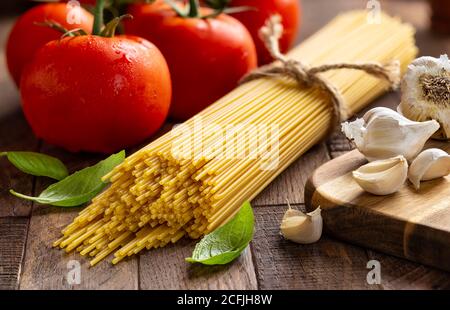 Spicchio di spaghetti crudi legato in un fascio con pomodori freschi di vite, aglio e basilico su un rustico tavolo di legno Foto Stock