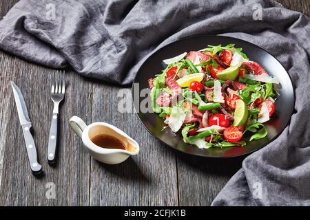 Insalata di bistecca di tagliata sirloin con fagioli verdi, rucola di pomodori ciliegini e parmigiano servita su un piatto nero su fondo di legno scuro con cu Foto Stock