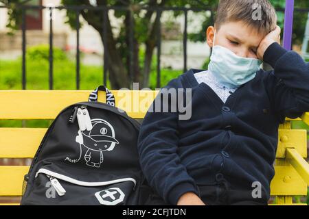 un ragazzo pensieroso in maschera pensare. un ragazzo triste che guarda seduto su una panchina con uno zaino nel cortile della scuola in attesa dell'autobus. Torna a schoo Foto Stock