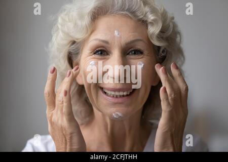 Head shot ritratto sorridente bella donna matura che applica la crema Foto Stock