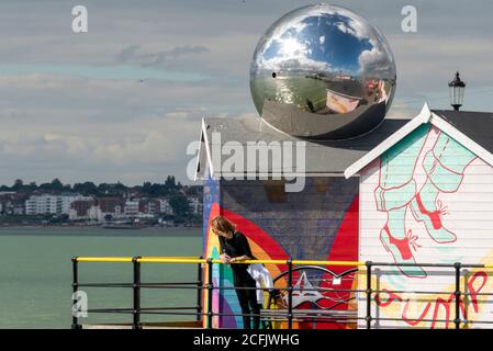 Southend on Sea, Essex, Regno Unito. 6 Settembre 2020. Il clima caldo e soleggiato ha attirato la gente verso la città balneare, con molti che prendono al molo. Una donna che pende sulle ringhiere del molo che si affacciano sul Tamigi con riflessi in un enorme globo di acciaio inossidabile Foto Stock