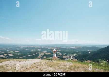 Vista posteriore di femmina in abito in piedi su collina con braccia sollevate e godere della libertà durante le vacanze Foto Stock