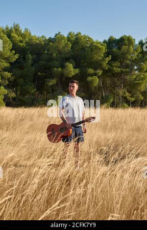 Musicista maschile pacifico con chitarra in caso di camminare lungo asciutto campo in estate e guardando via Foto Stock