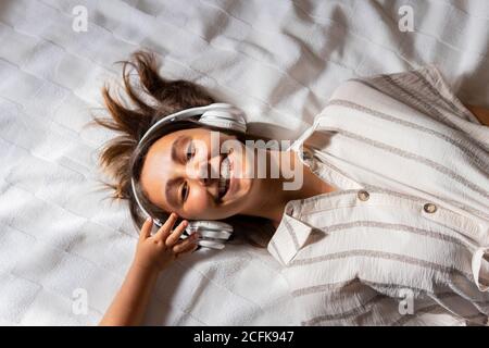 Ritaglia il bambino toccando le cuffie di Donna sdraiata sul letto in camera da letto e godendo di ascoltare la musica mentre guarda una macchina fotografica Foto Stock