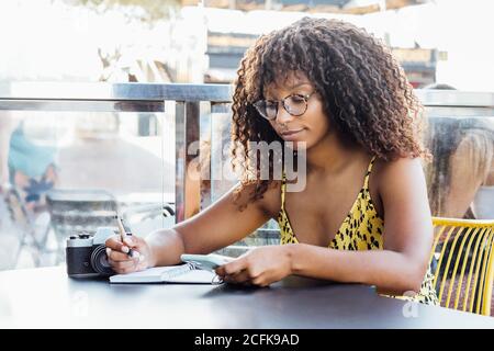 Nero lavoratore autonomo Donna seduta a tavola in caffè e lavorare a distanza mentre si scrive in blocco note Foto Stock