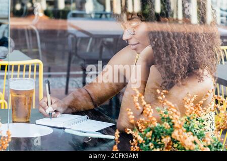 Vista laterale di nero lavoratore autonomo Donna seduta al tavolo in caffè e lavorare a distanza mentre si scrive in blocco note Foto Stock