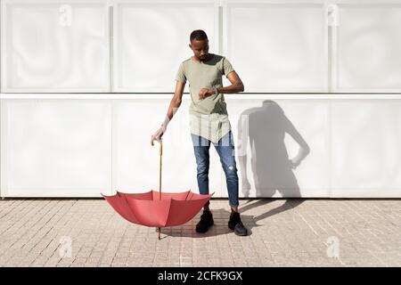 Serio uomo afroamericano che tiene un ombrello capovolto sulla strada il giorno di sole e controllare l'orario sul orologio da polso Foto Stock