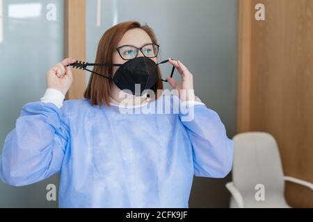 Operatrice medica femminile in uniforme blu e occhiali che indossano maschera protettiva nera con cinturino regolabile mentre si lavora in stile moderno clinica durante la pandemia del coronavirus Foto Stock