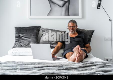Sorridente imprenditore maschile di mezza età seduto a letto e digitando su un computer portatile mentre si lavora in remoto su un progetto online Foto Stock
