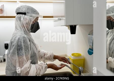 Vista laterale del medico femminile in tuta protettiva e viso proteggi con la maschera lavando le mani durante la preparazione per la procedura medica in clinica moderna Foto Stock