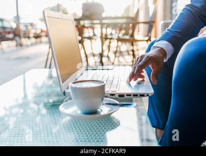 Vista laterale del raccolto anonimo uomo d'affari nero in tuta formale seduto a tavola con una tazza di caffè e digitando computer portatile mentre si lavora in un bar Foto Stock