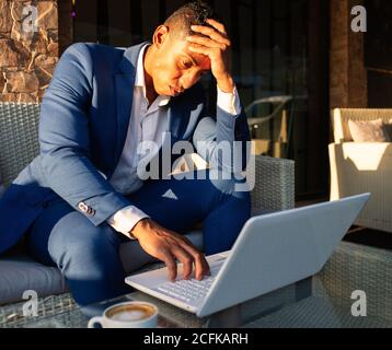 Giovane imprenditore africano americano maschio in abbigliamento formale su cui lavorare computer portatile e pensare a problemi finanziari mentre si siede in sala zona di caffetteria moderna Foto Stock