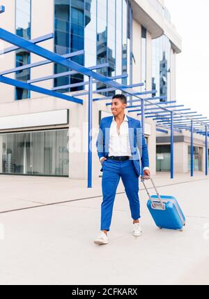 Lunghezza completa di giovane imprenditore etnico maschile fiducioso in stile vestito con valigia a piedi vicino a un edificio moderno Foto Stock