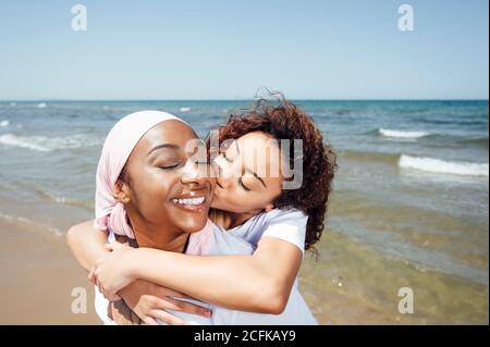 Felice madre nera piggybacking giovane figlia mentre si divertono con occhi chiusi al mare in estate Foto Stock