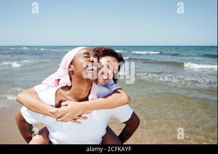 Felice madre nera piggybacking giovane figlia mentre si divertono con occhi chiusi al mare in estate Foto Stock