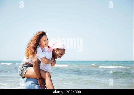 Felice madre nera piggybacking giovane figlia mentre si divertono con occhi chiusi al mare in estate Foto Stock