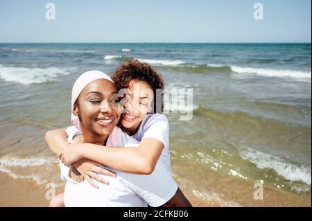 Felice madre nera piggybacking giovane figlia mentre si divertono con occhi chiusi al mare in estate Foto Stock