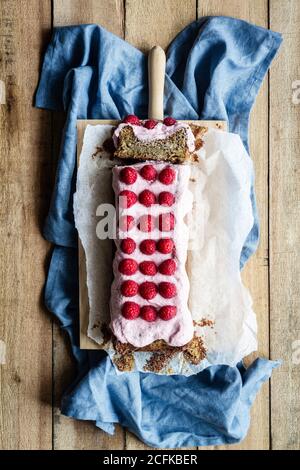 Vista dall'alto di una deliziosa dolce torta fatta in casa sormontata da una frusta crema e guarnita con lamponi freschi su carta da forno a tavola con ingredienti in cucina Foto Stock