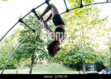 Vista laterale ad angolo basso della giovane atletica afroamericana in abbigliamento sportivo appeso capovolto sulla scala ad arco durante l'esercizio nel parco estivo Foto Stock