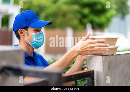 Asian consegnare l'uomo con maschera facciale in camicia blu maneggiando scatole di cibo e messo sul posto recinzione della casa del cliente come consegna di cibo senza contatto. Questo c Foto Stock