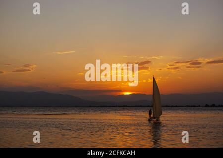 Piccola barca a vela in mare al tramonto con il sole e le nuvole sullo sfondo. Vela in un'avventura estiva nel Mar Mediterraneo Foto Stock