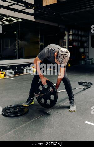 Atleta maschile in abbigliamento sportivo mettendo piatti pesanti sul barbell mentre preparazione per l'allenamento di sollevamento pesi in palestra Foto Stock