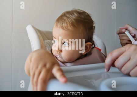 Bambina che guarda da parte seduta distrattamente su una sedia alta con un bib mentre è alimentato da persona irriconoscibile con un cucchiaio Foto Stock