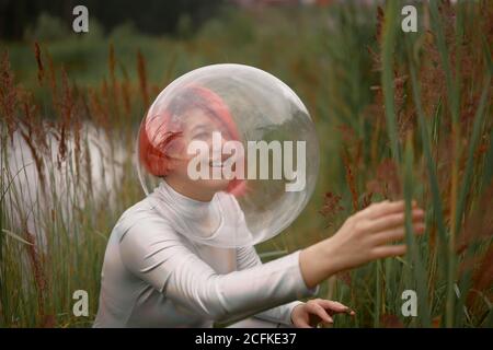 Vista laterale di felice futurista giovane capelli rossi femmina in tuta spaziale in argento e casco in vetro in piedi vicino a piante verdi sul fiume che allagano Foto Stock