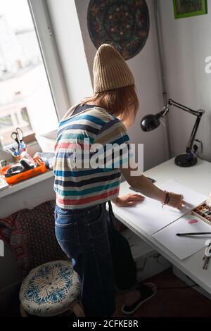 Vista laterale ad alto angolo di una artista femminile concentrata in piedi tavolo in officina moderna e uso acquerello per la pittura Foto Stock