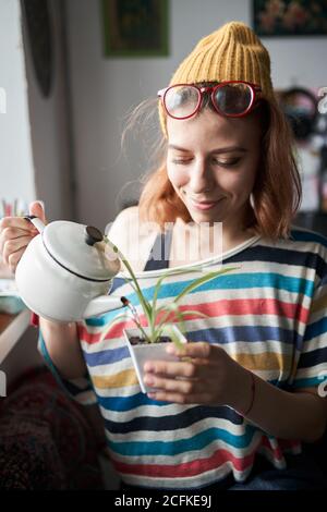 Contenuto femminile in abiti fantasia in piedi in soggiorno e. annaffiatoio in vaso Foto Stock