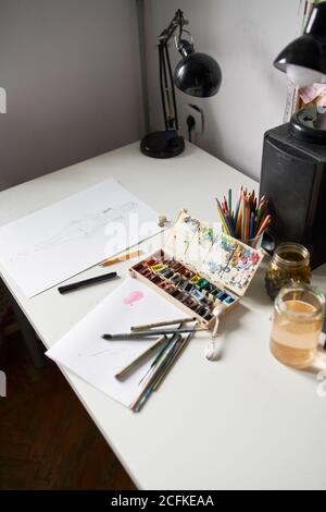 Angolo elevato di vari pennelli e palette di acquerelli disposti sopra tavolo con carta e vasetto di vetro d'acqua in arte studio Foto Stock