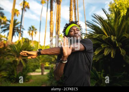 Giovane atleta afro-americano di fiducia in abbigliamento sportivo alla moda stretching armi mentre si allenano nel parco con alberi tropicali verdi Foto Stock