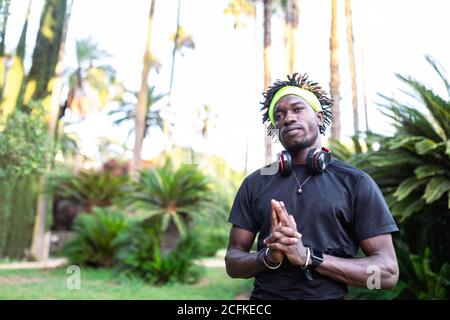 Giovane atleta africano-americano maschio in abbigliamento sportivo con cuffie collo che tiene le mani aggrappate e che guarda lontano pensieroso mentre si è in piedi in verde parco tropicale Foto Stock