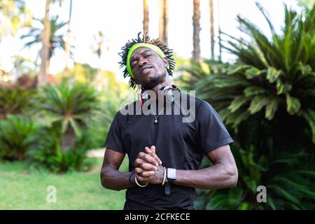 Giovane atleta africano-americano maschio in abbigliamento sportivo con cuffie collo che tiene le mani aggrappate mentre si trova in piedi nel verde parco tropicale Foto Stock